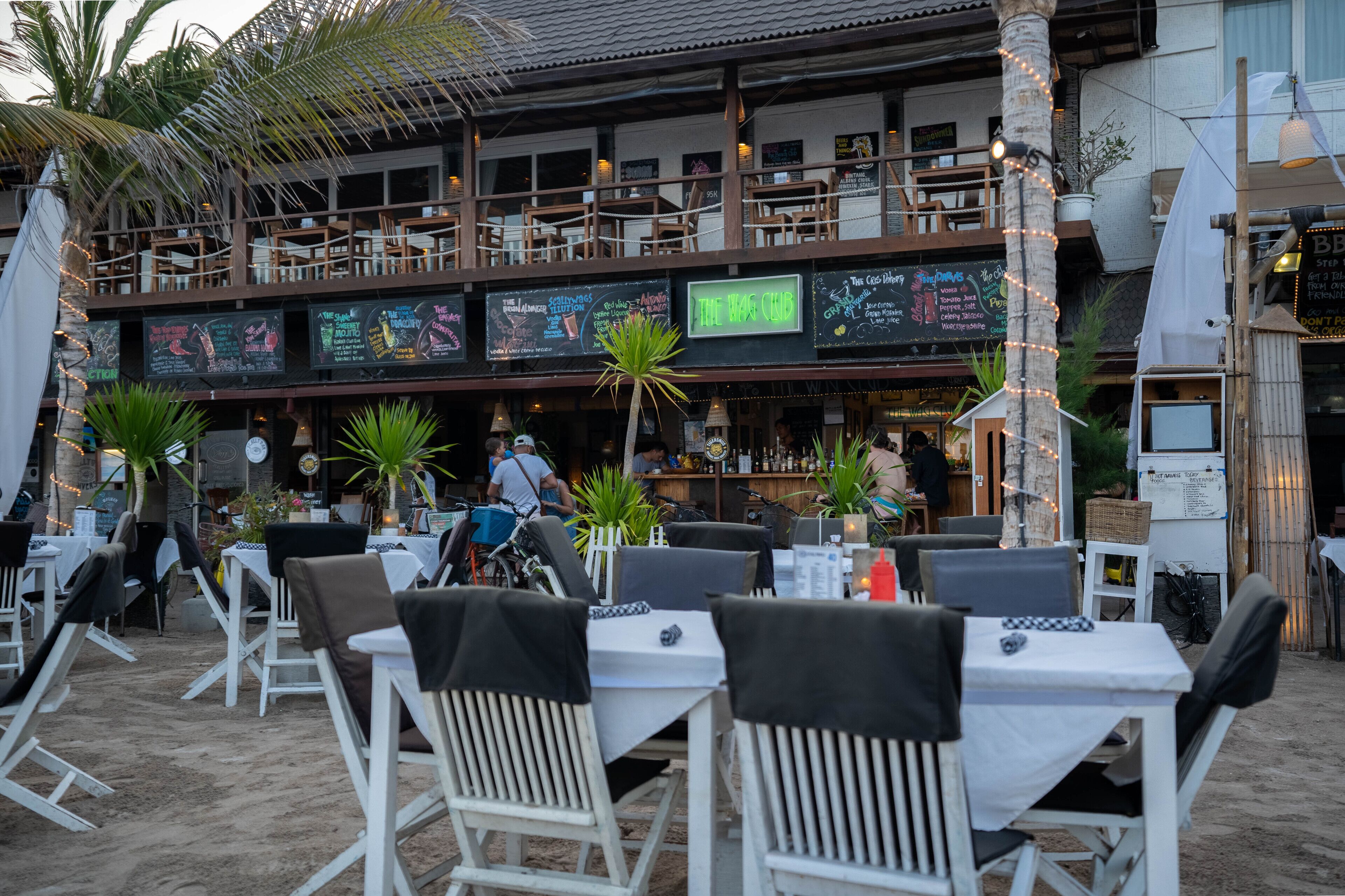 An outdoor restaurant with white tables and chairs, a neon sign reading "The Bar Club", and a balcony with wooden chairs.