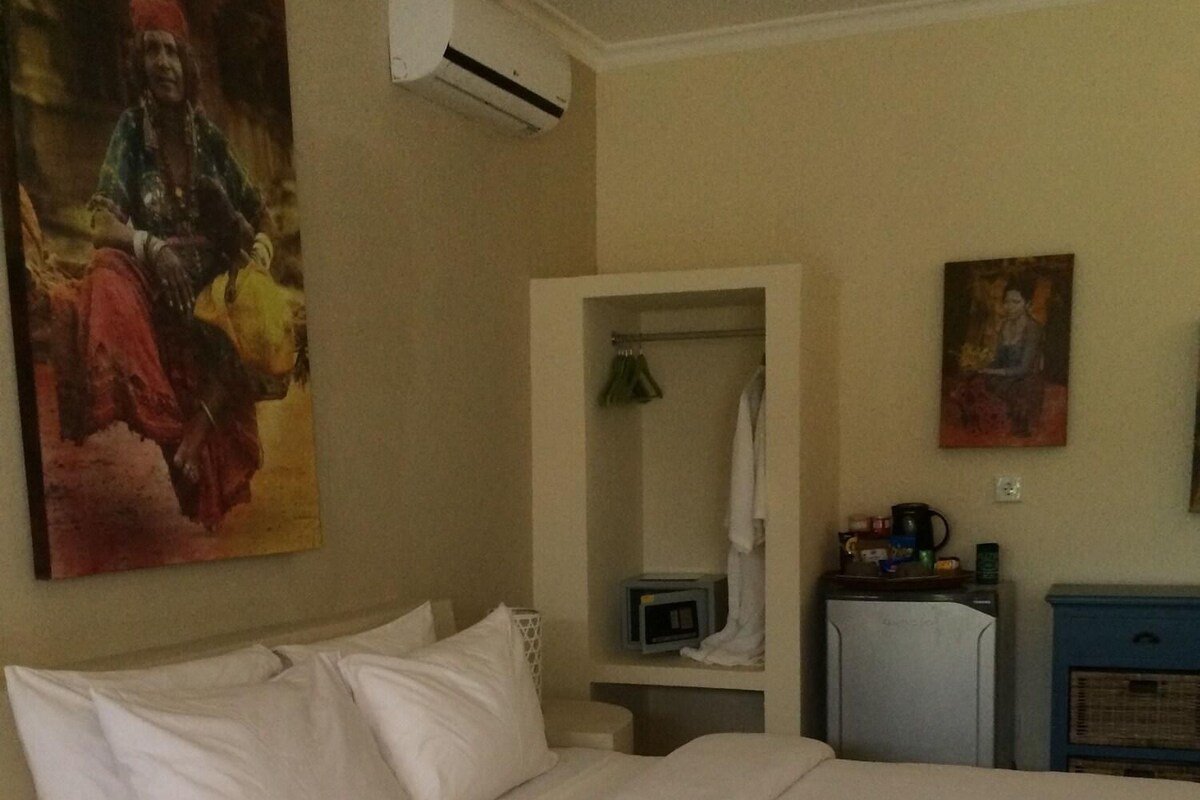 A hotel room with a large bed, an air conditioning unit, a small desk with a lamp, and a painting on the wall.