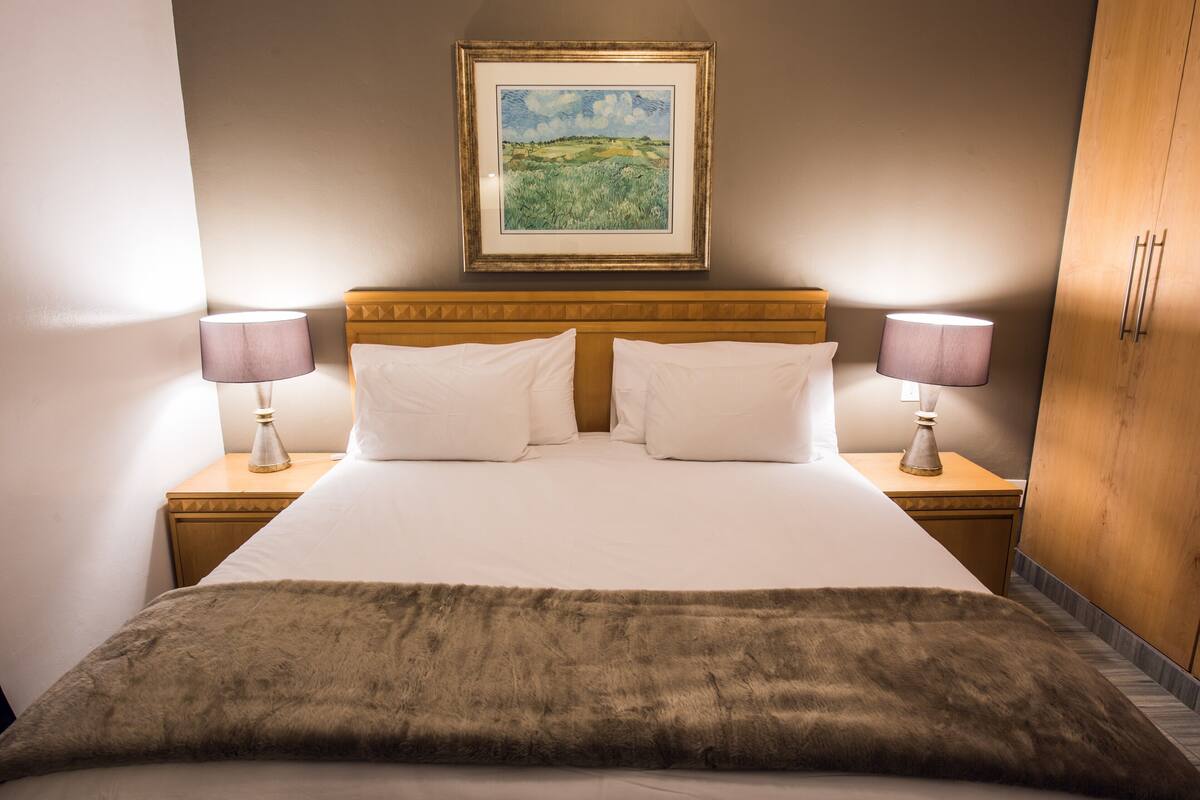 Una cama bien hecha con sábanas blancas y dos almohadas, flanqueada por dos mesitas de noche con lámparas. Un cuadro de paisaje enmarcados decora la pared sobre la cama.