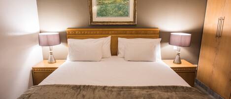 A neatly made bed with white linens and two pillows, flanked by two bedside tables with lamps. A framed landscape painting hangs above the bed.