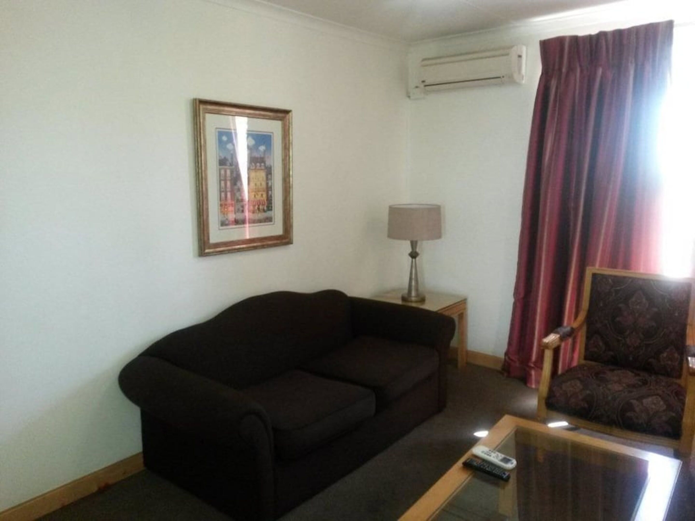 Uma sala de estar com um sofá escuro, uma mesinha com abajur, uma cadeira e um quadro na parede.