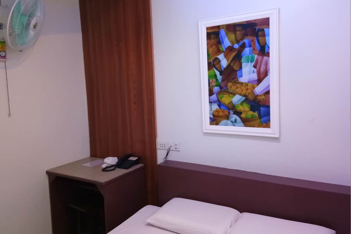 A hotel room with a bed, a small desk with a phone, a wall-mounted fan, and a framed abstract painting.