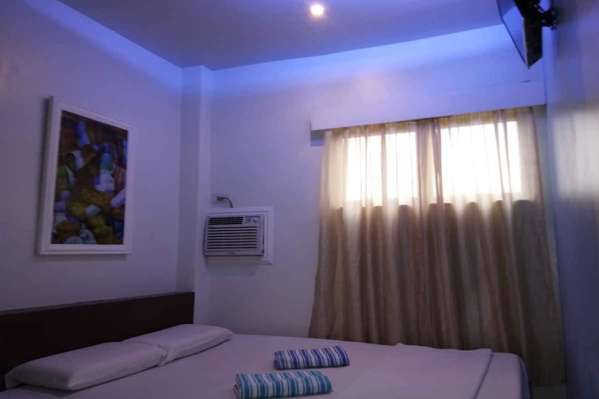 A hotel room with a bed, pillows, a painting on the wall, an air conditioning unit, and a window with curtains.