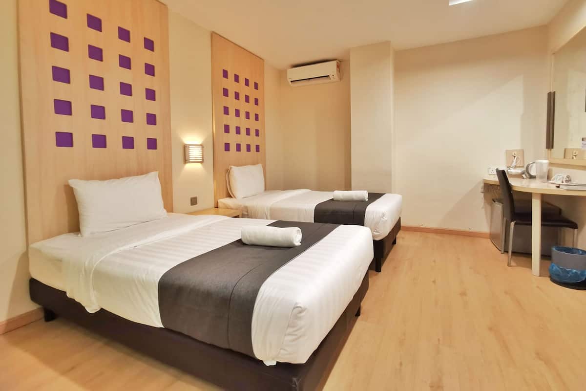 Ein Hotelzimmer mit zwei Betten, einem Schreibtisch und einem Stuhl. Es gibt eine Wand-Klimaanlage und eine Deckenleuchte.