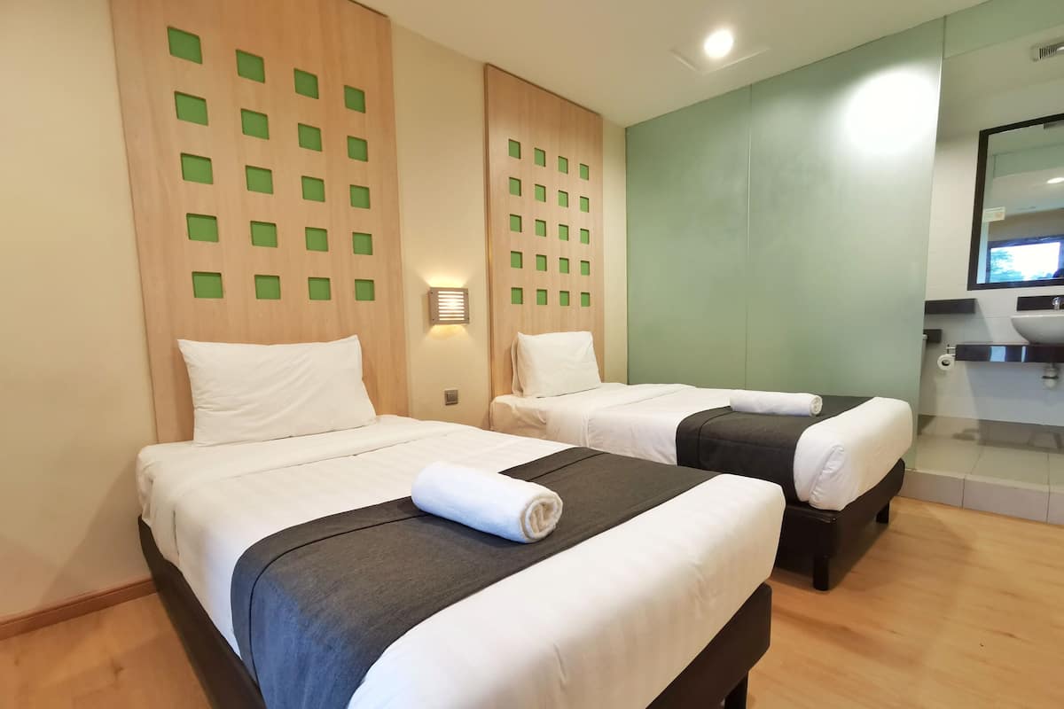 Ein Hotelzimmer mit zwei Betten, einem Holzkopfteil mit grünen quadratischen Ausschnitten, einer Wandleuchte und einem Badezimmer mit Waschbecken und Spiegel.