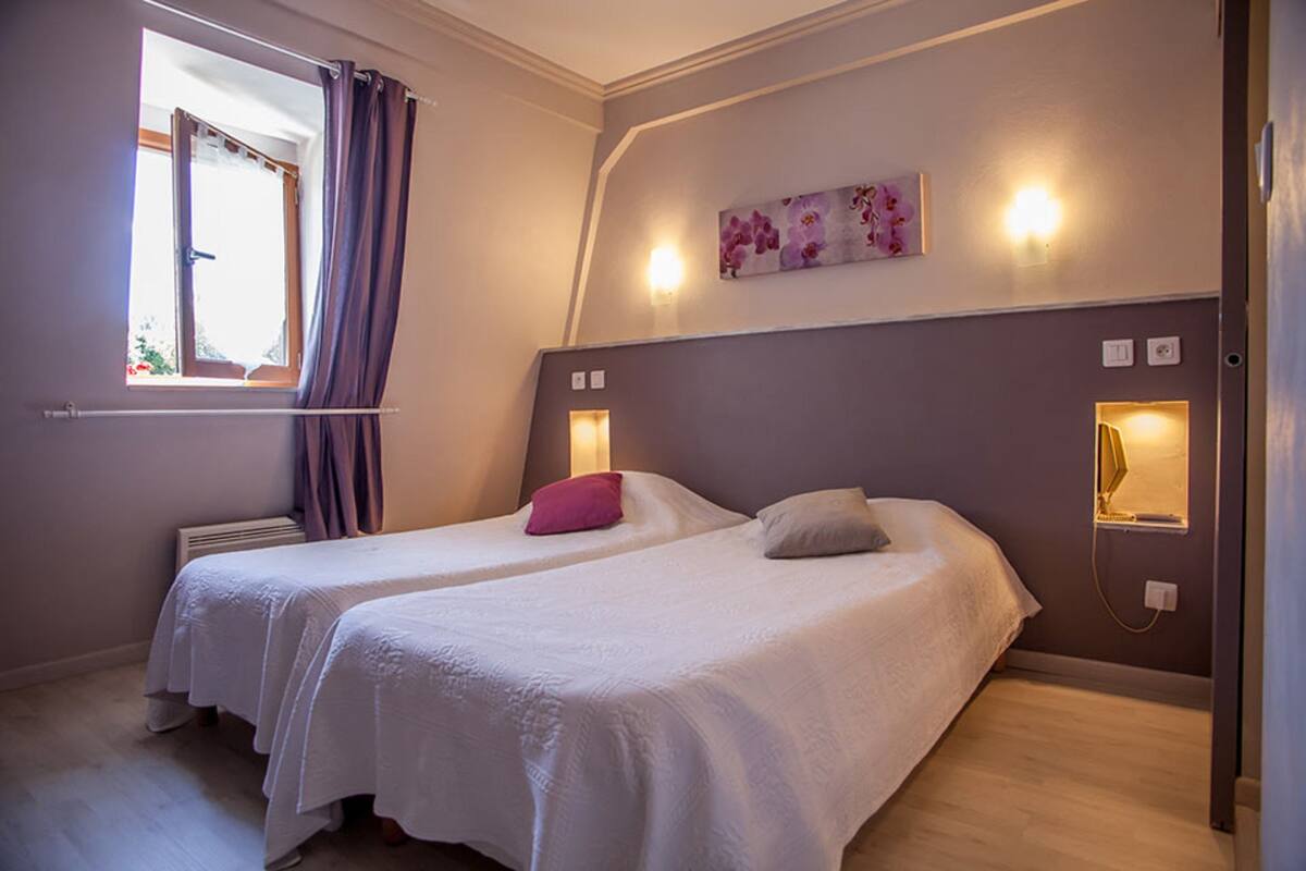 A bedroom with two beds, a window with curtains, a wall-mounted light, and a floral painting.