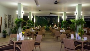 Ein Restaurant mit Glastischen und Korbstühlen, umgeben von Topfpflanzen und gerahmten Kunstwerken an den Wänden.