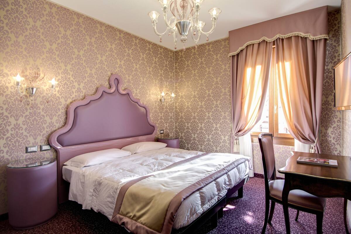 A bedroom with a bed, bedside tables, a chair, a desk, and a chandelier.