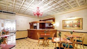 A hotel lobby with a classic design, featuring a central reception desk, multiple seating areas with tables and chairs, and a decorative ceiling with a chandelier.