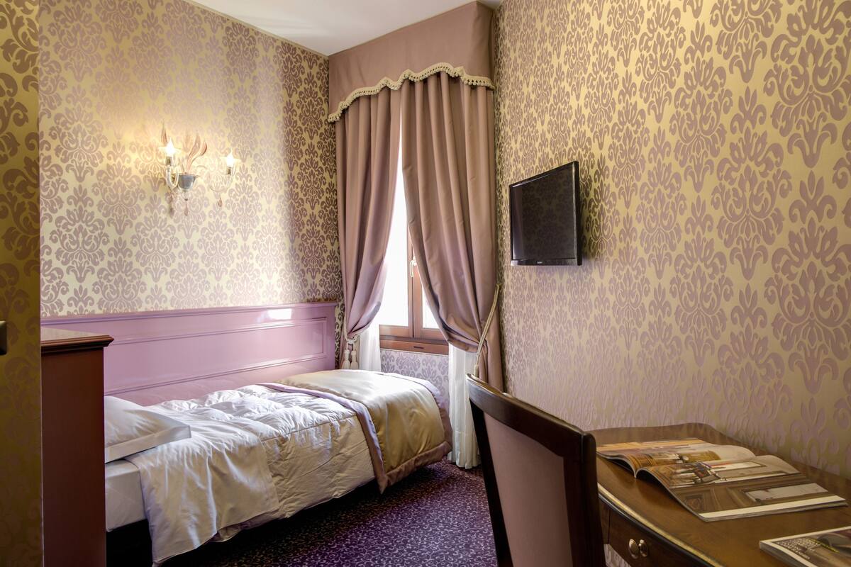 A hotel room with a bed, a desk, a TV, and patterned wallpaper.