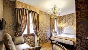 A hotel room with a bed, a sofa, a chair, a chandelier, and patterned wallpaper.