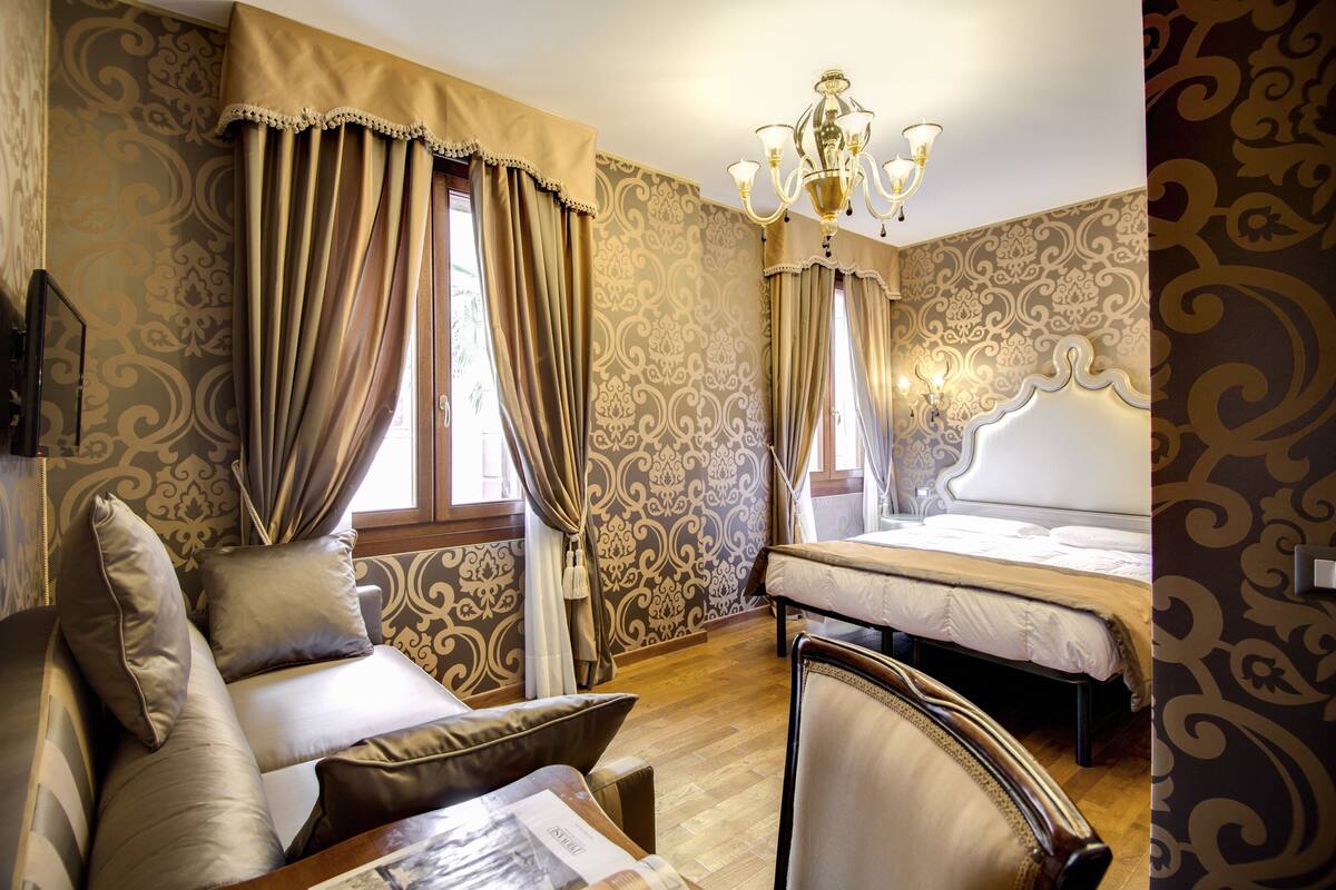 A hotel room with a bed, a sofa, a chair, a chandelier, and patterned wallpaper.