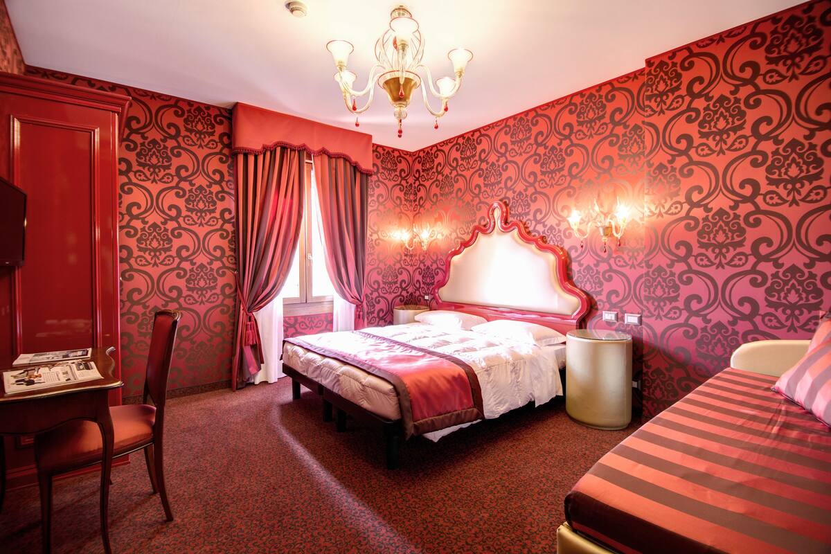 A hotel room with a large bed, a desk with a chair, a chandelier, and patterned wallpaper.