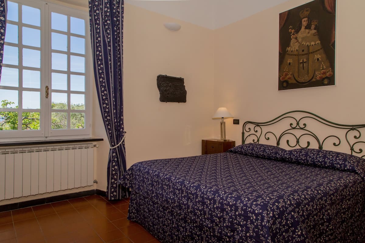 Une chambre dotée d’une grande fenêtre, d’un lit recouvert d’une couvre-lit à motifs floraux, d’une table de chevet avec une lampe et d’une œuvre d’art accrochée au mur.