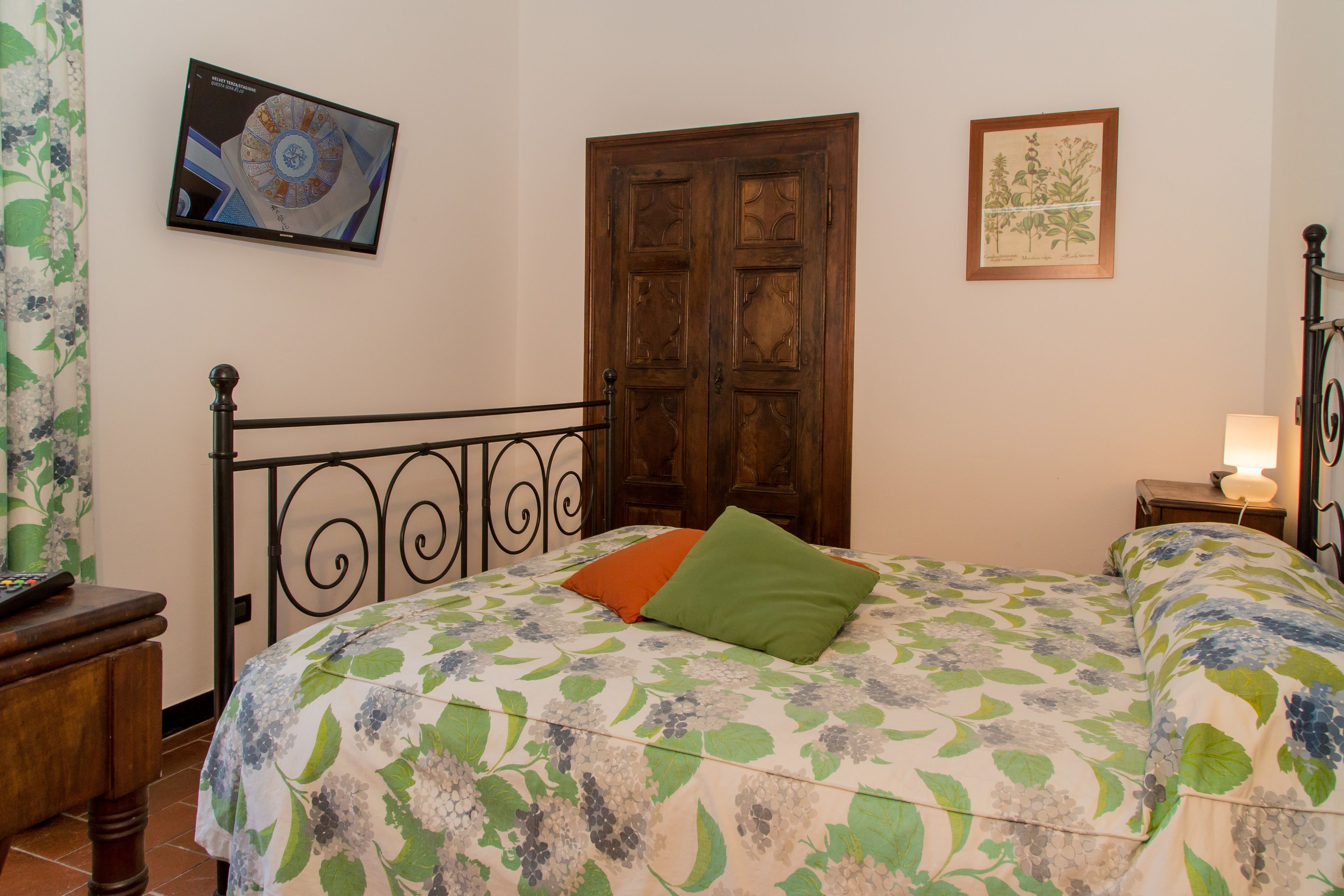 Une chambre à coucher avec un lit, une porte en bois, un téléviseur fixé au mur et une illustration botanique encadrée.
