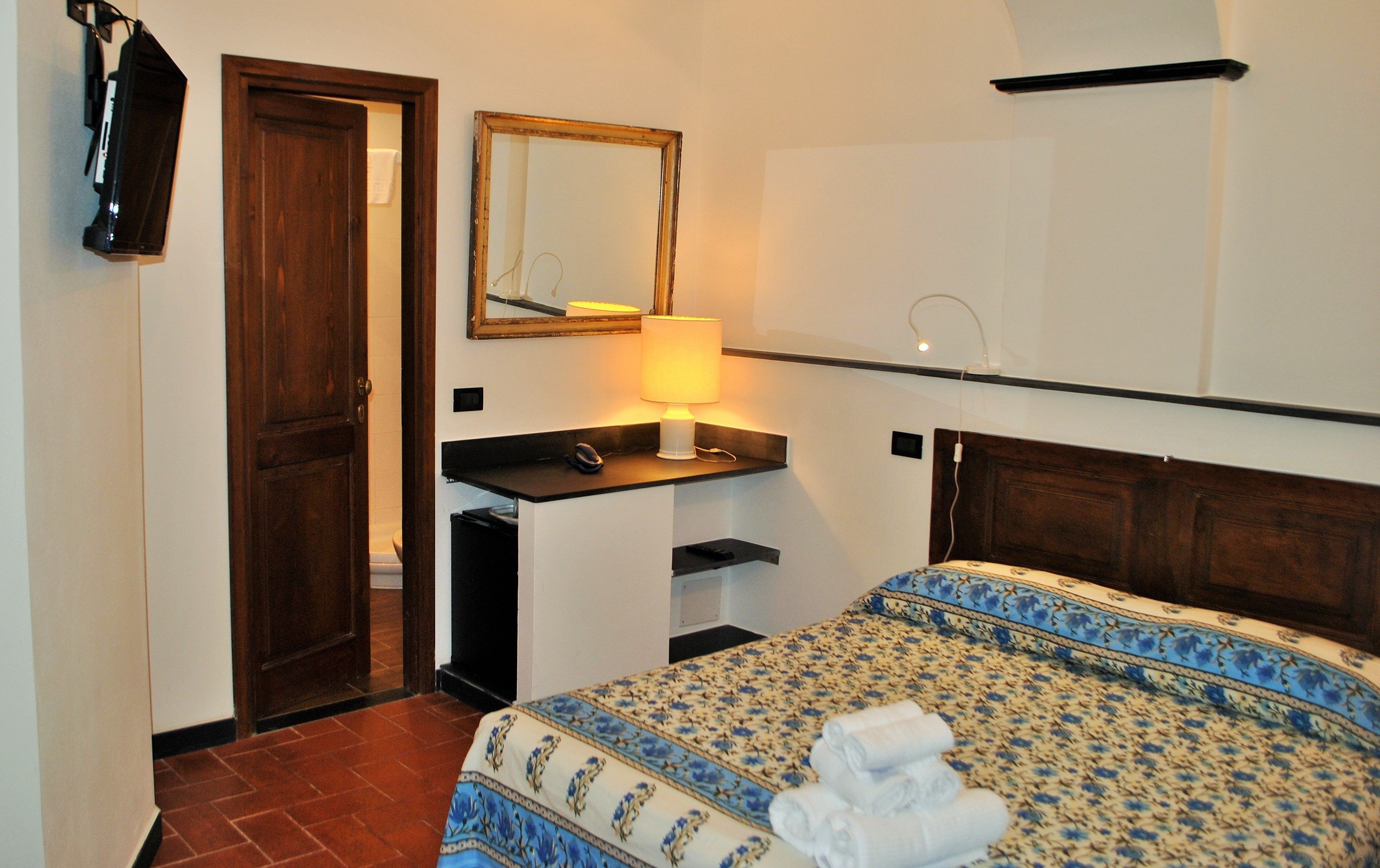 A hotel room with a bed, a desk with a lamp, a mirror, and a door.