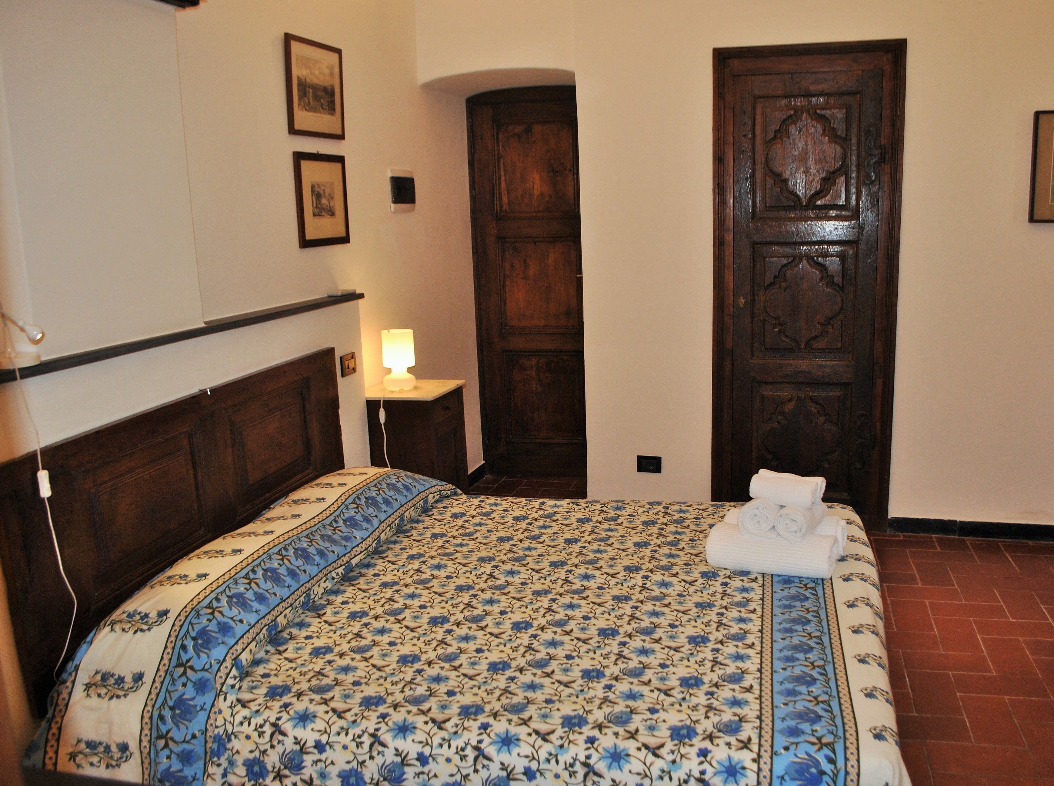 A bedroom with a bed, a wooden door, a nightstand with a lamp, and two framed pictures on the wall.