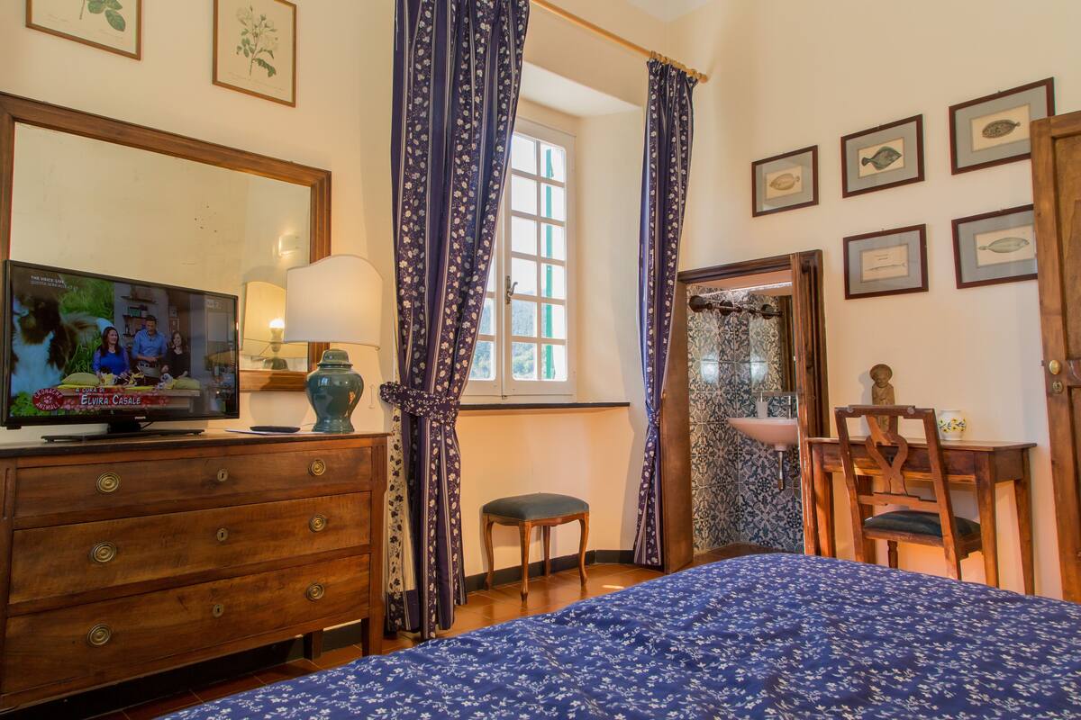 Une chambre à coucher comprenant un lit, une commode, un téléviseur, une fenêtre avec des rideaux, une chaise et des tableaux encadrés accrochés au mur.