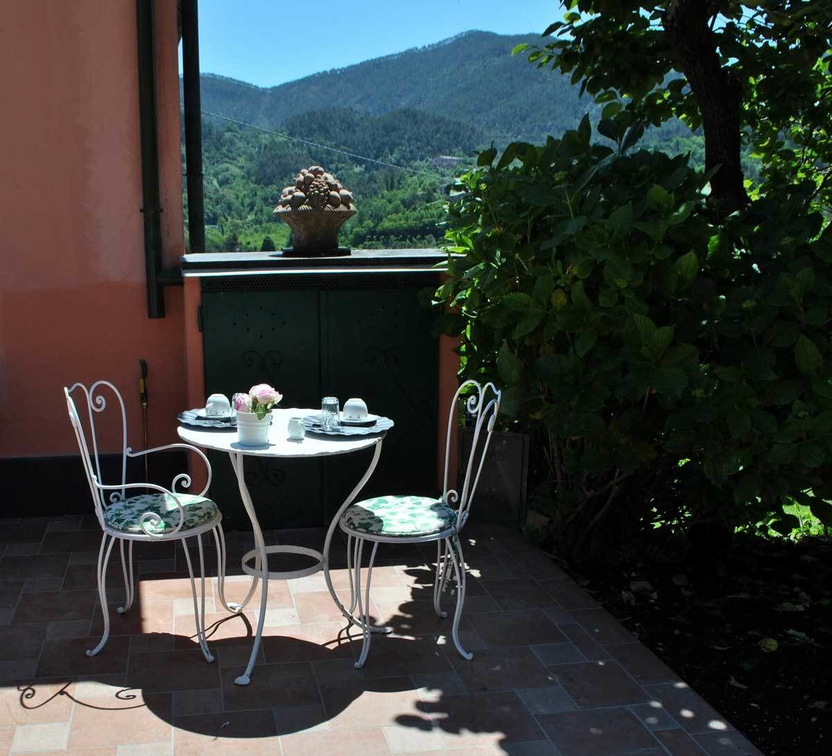 Patio esterno con tavolo rotondo e due sedie, una pianta in vaso e vista su una montagna.