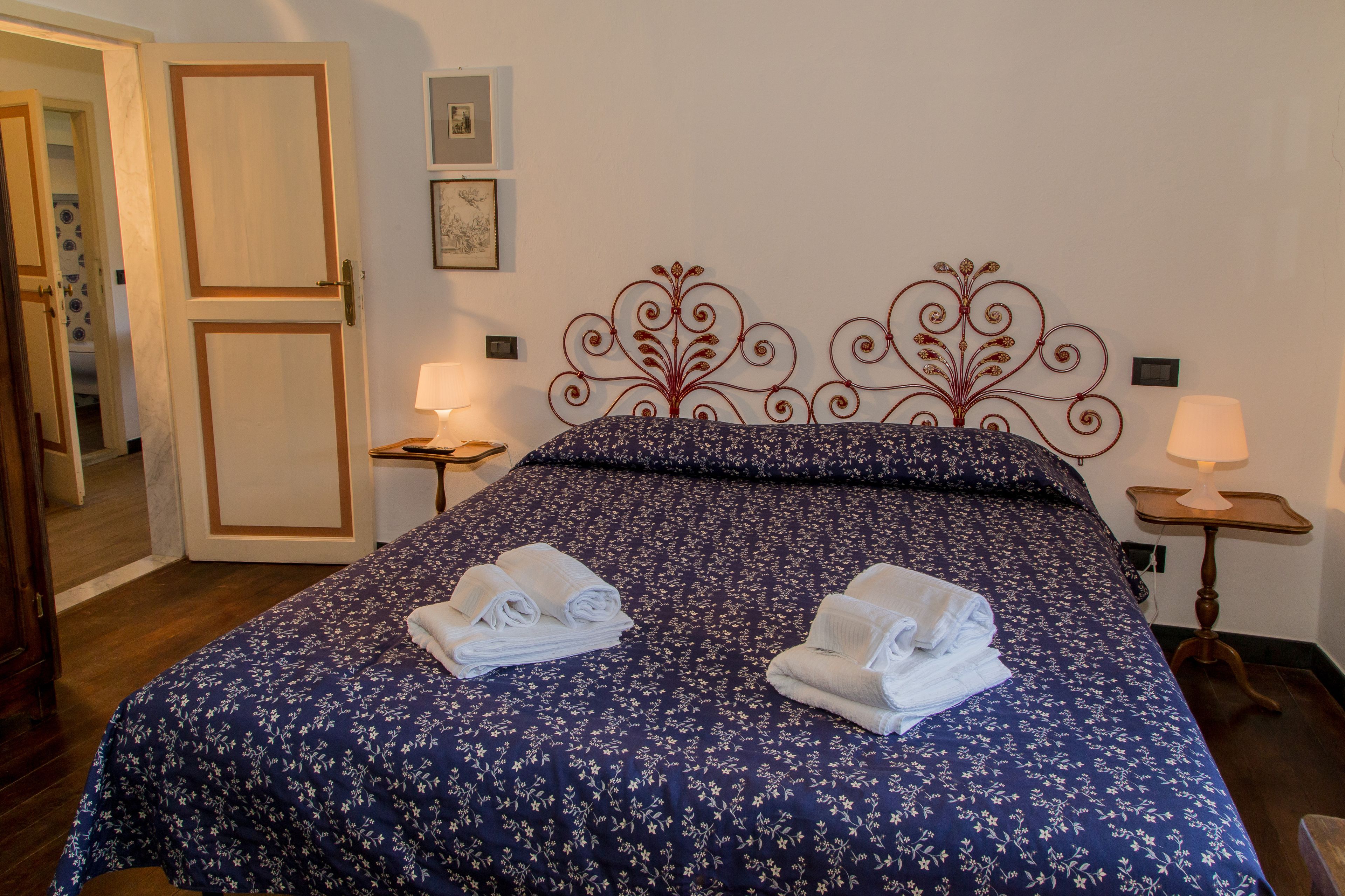 Un lit bien fait, recouvert d’une couvre-lit à motifs floraux, avec deux serviettes enroulées posées sur le lit et une tête de lit décorative en métal.