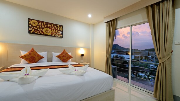 A hotel room with a large bed, two pillows, a painting on the wall, and a view of a cityscape through a large window. - Baan Kure Ngern Aonang (Krabi)