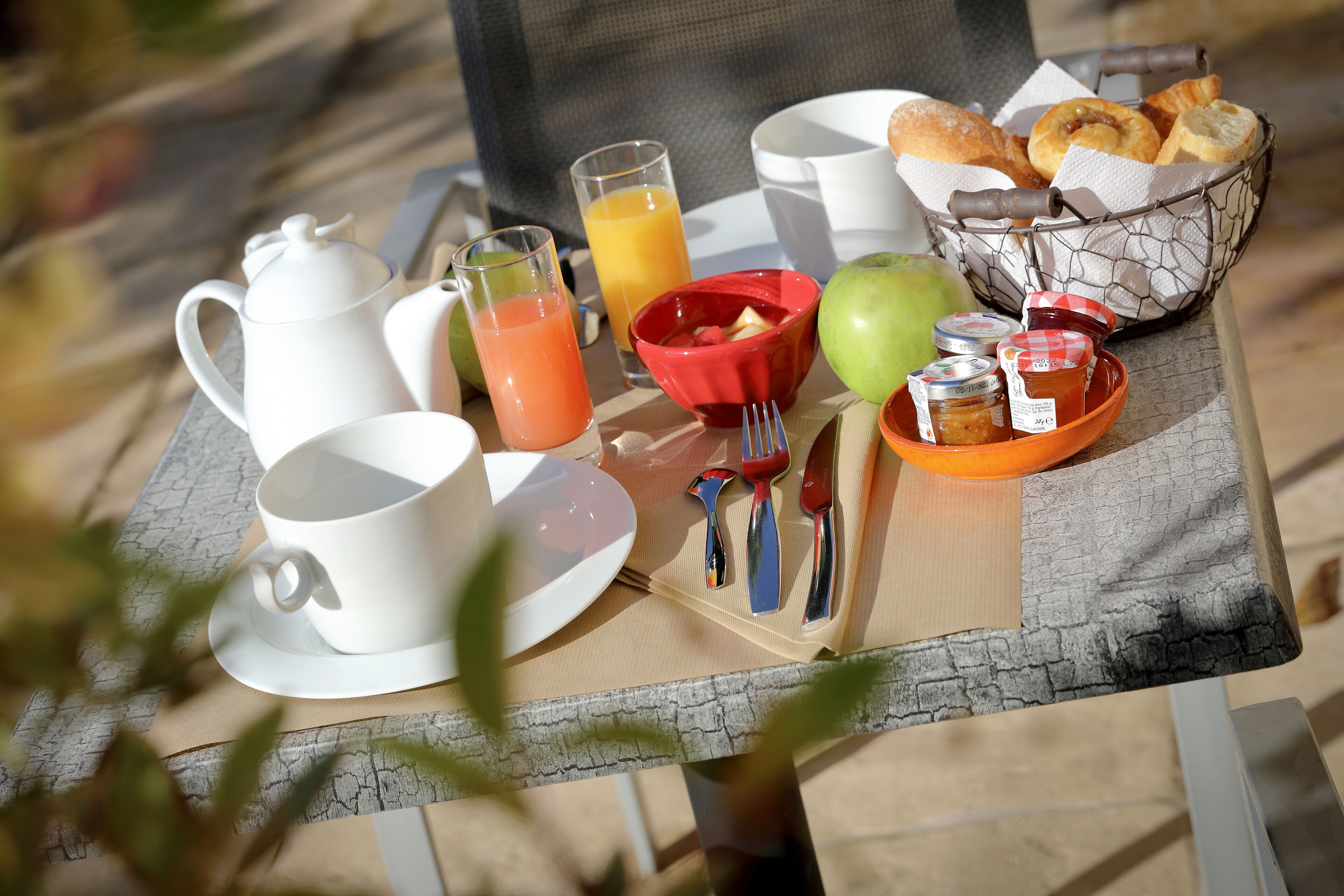 Een ontbijttafel met een witte theepot, kopjes, een rode kom, een mand met brood en een dienblad met fruit en toevoegingen.