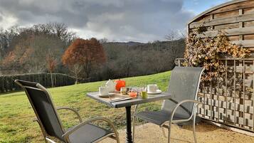 Un espace extérieur aménagé avec deux chaises et une table, une clôture en bois et une vue sur un champ verdoyant et des arbres sous un ciel nuageux.