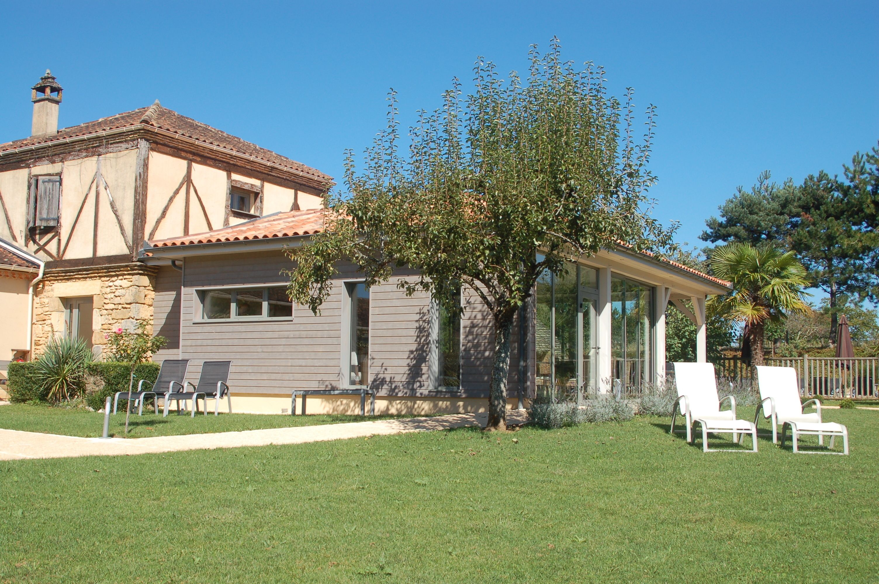 A two-story house with a stone facade, a tiled roof, and a large tree in the front yard. There are two white lounge chairs and a pathway leading to the house.