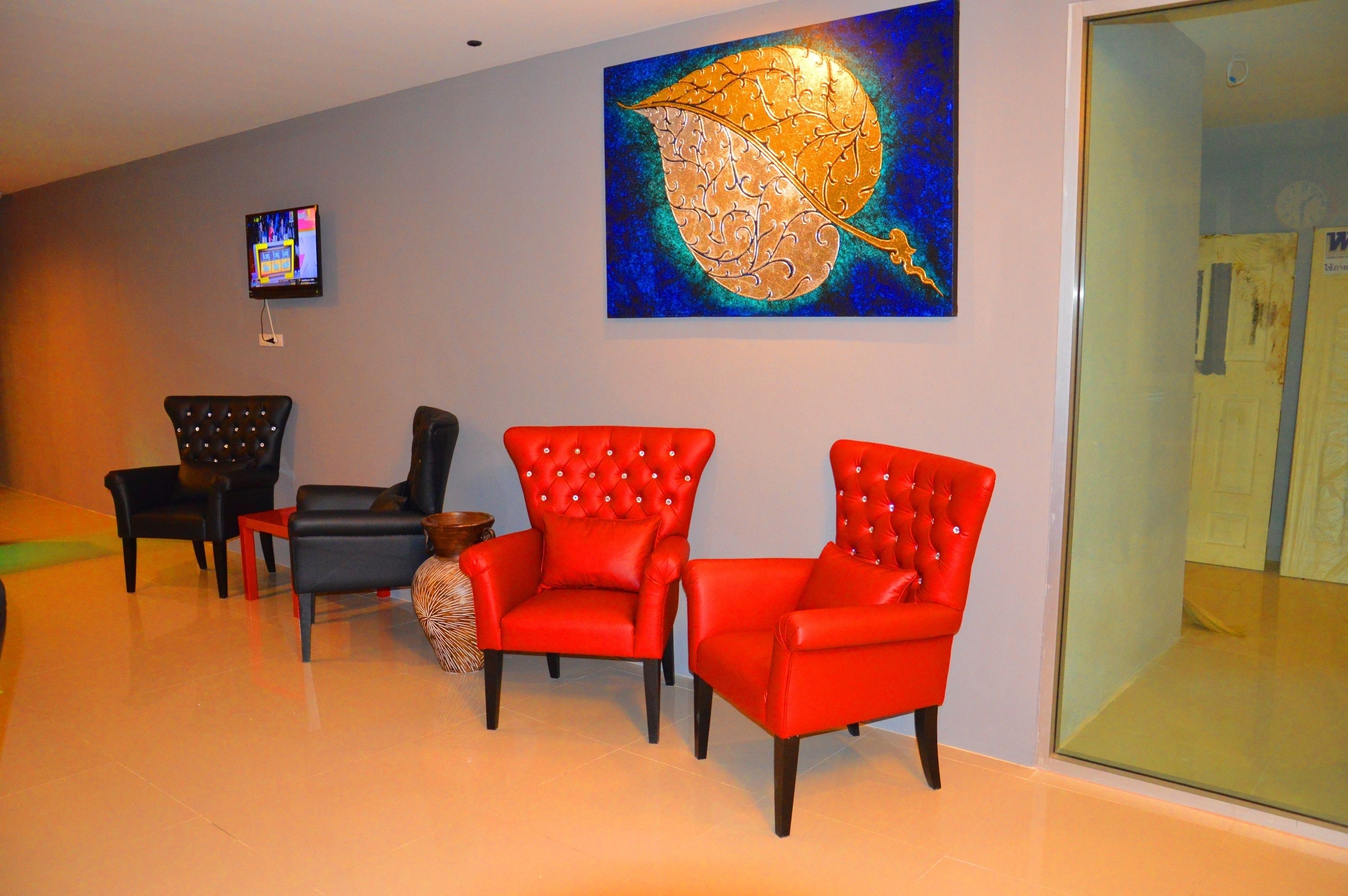A room with three red chairs, a black chair, and a small table. There is a painting on the wall and a television mounted on the wall.