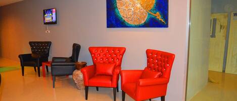 A room with three red chairs, a black chair, and a small table. There is a painting on the wall and a television mounted on the wall.