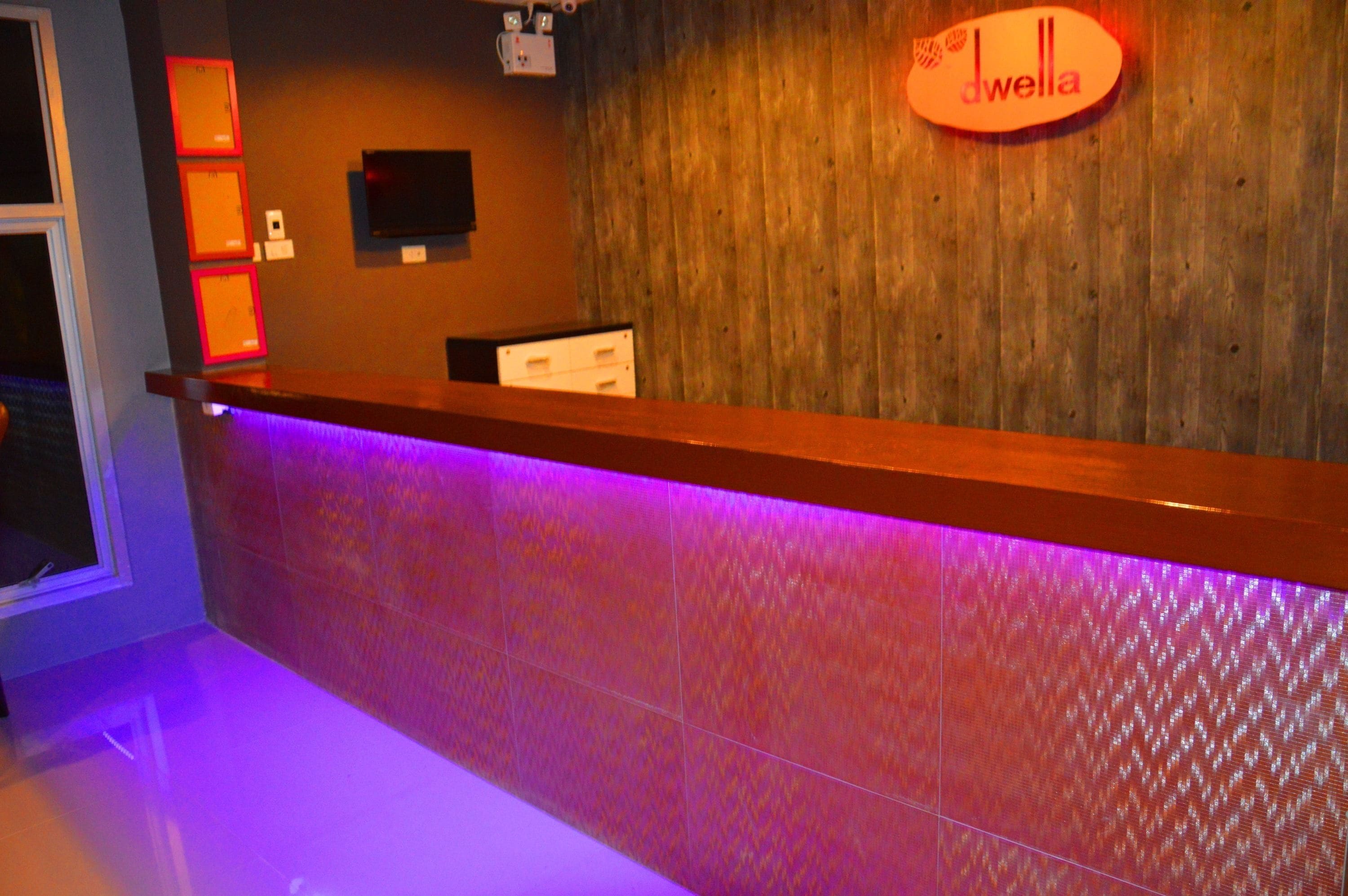 A reception area with a wooden wall, a 'dwella' sign, a flat-screen TV, and a purple-lit reception desk.