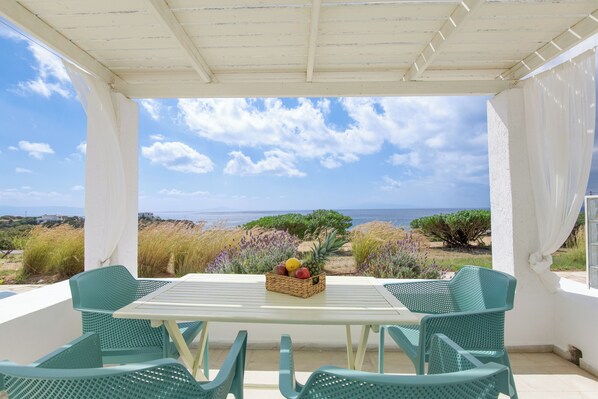 A covered outdoor seating area with a table and chairs, a fruit basket on the table, and a view of the sea and landscape. - Glyfa Village  (Paros)