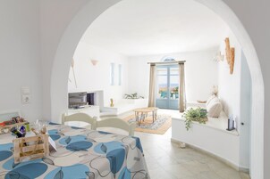 Un interior luminoso y blanco con zona de comedor que incluye una mesa con mantel azul y blanco, sillas y una mesita auxiliar con una botella y vasos.