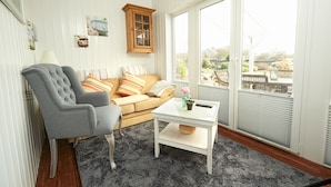 A living room with a grey armchair, a beige sofa with striped pillows, a white coffee table with a vase and remote control, and large windows with blinds. - Hotel Fjordkroen (Tappernoje)