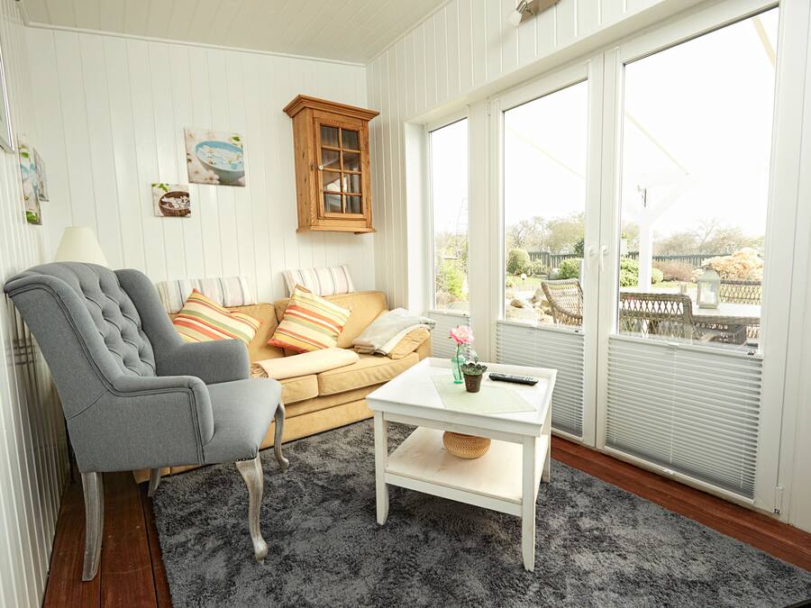 A living room with a grey armchair, a beige sofa with striped pillows, a white coffee table with a vase and remote control, and large windows with blinds.