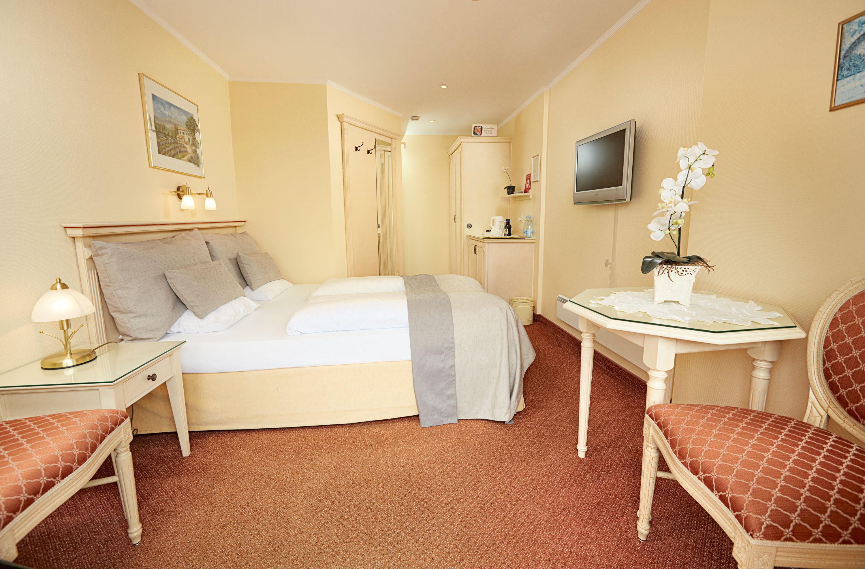 A hotel room with a bed, bedside table, lamp, chair, TV, and a small table with a vase of flowers.