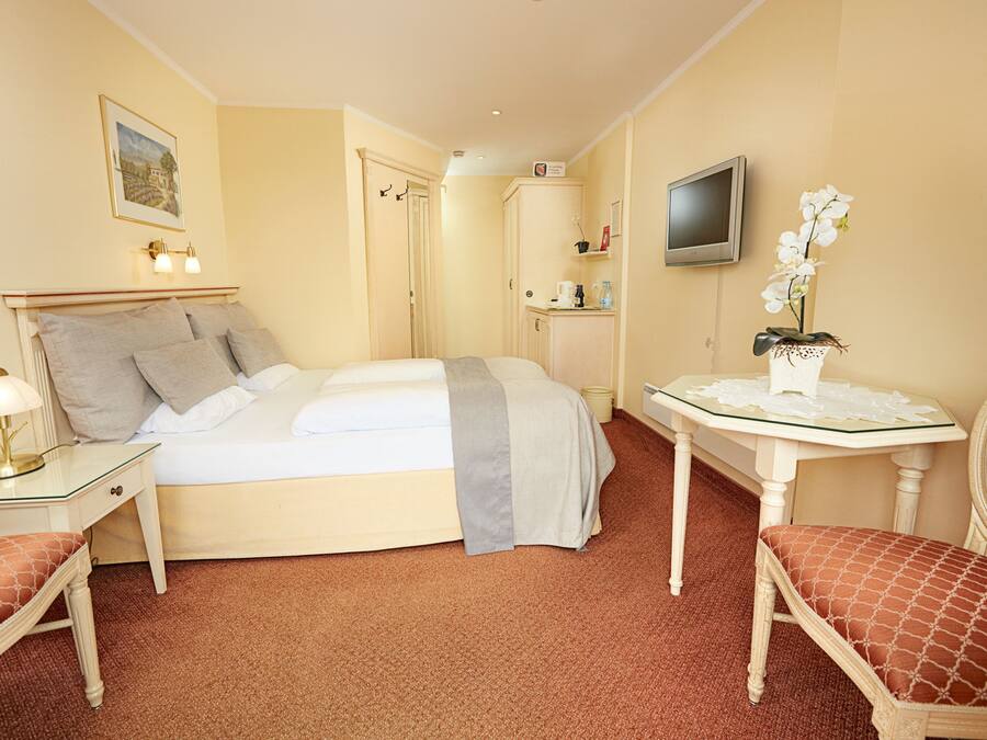 A hotel room with a bed, bedside table, lamp, chair, TV, and a small table with a vase of flowers.