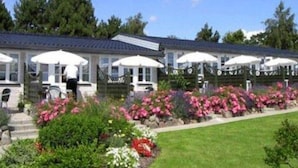 A row of single-story buildings with covered outdoor seating areas, surrounded by a well-maintained garden with various flowering plants. - Hotel Fjordkroen (Tappernoje)