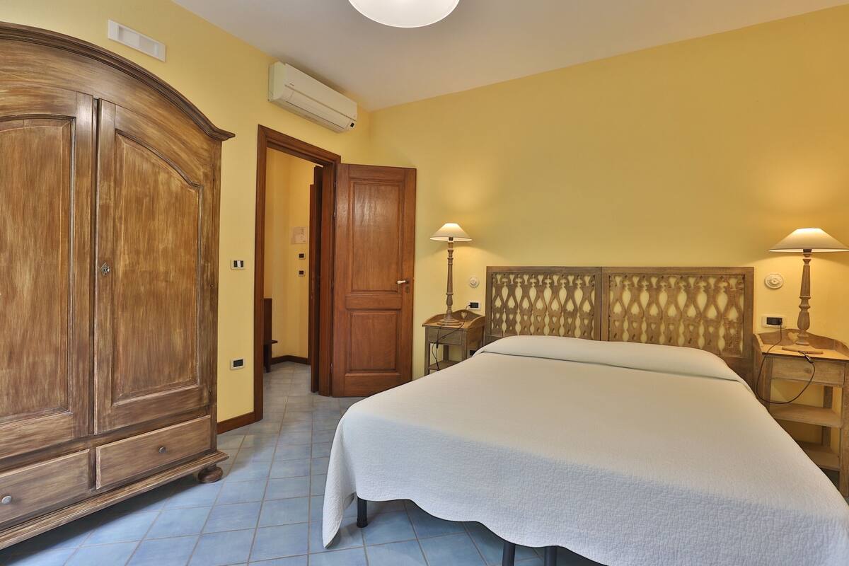 A bedroom with a large wooden wardrobe, a bed with a white bedspread, two bedside lamps, and a headboard with a decorative pattern.