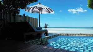 A poolside area with a wooden deck, a lounge chair, a striped umbrella, and a view of the beach. - Casa De Mar (Koh Samui)