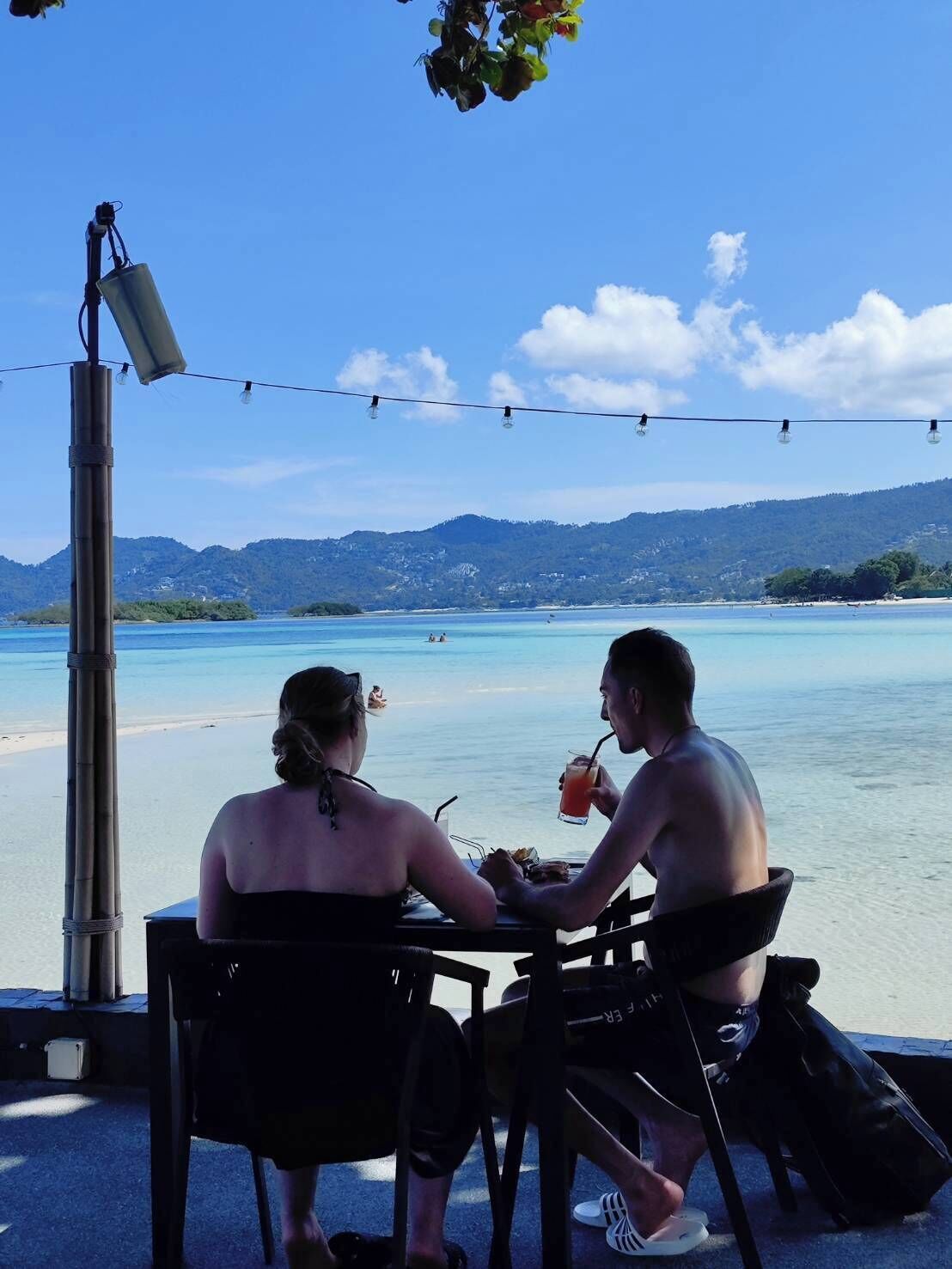 Dos personas cenando en una mesa al aire libre con vistas a una playa y montañas.