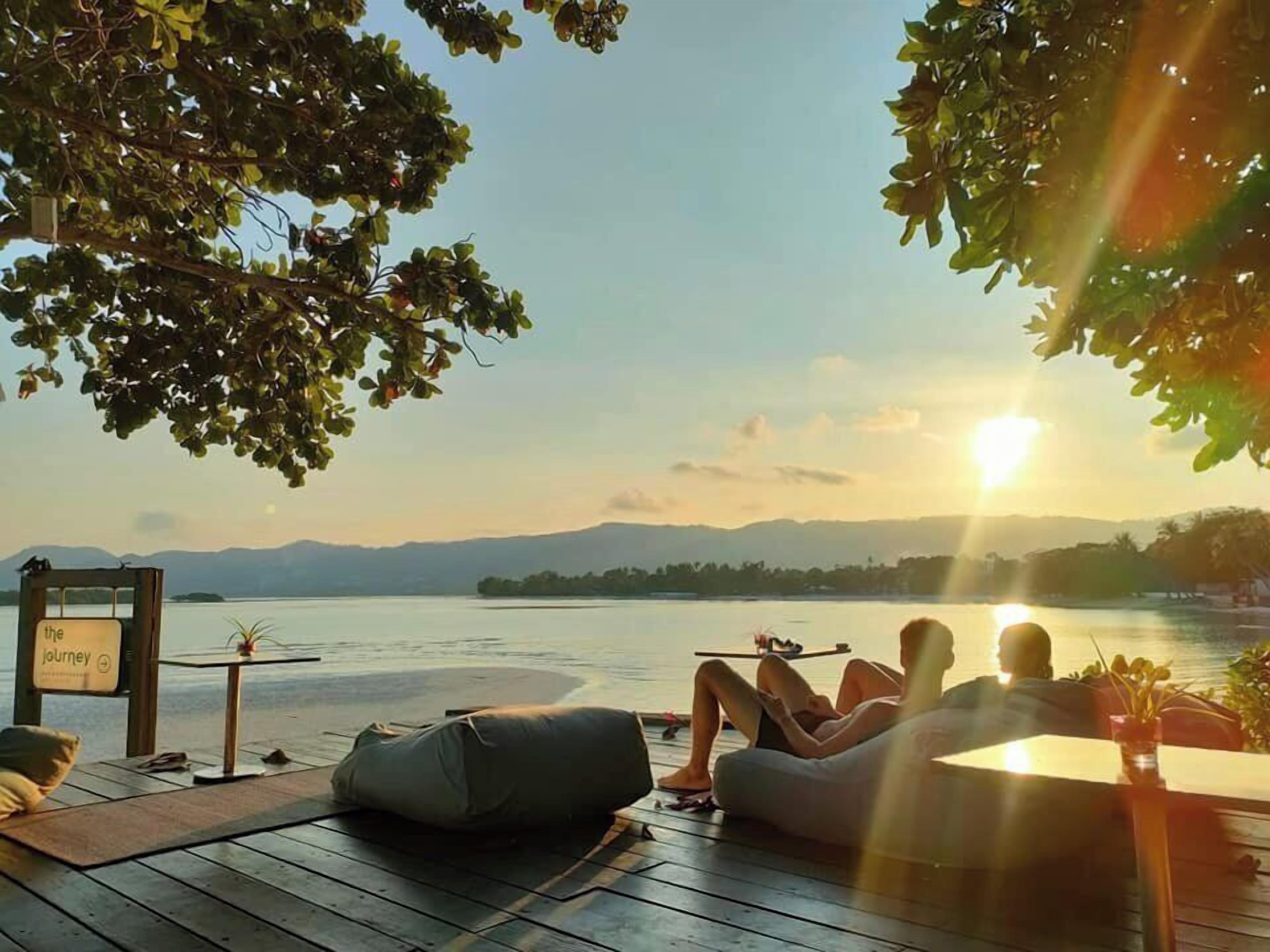 Um deck com vista para um lago e montanhas, uma placa com a frase 'a jornada' e uma pessoa relaxando.