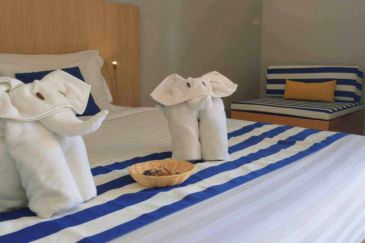 A hotel room with a large bed featuring blue and white striped bedding, two towels shaped like elephants, a small wicker basket, and a bedside table with a lamp.