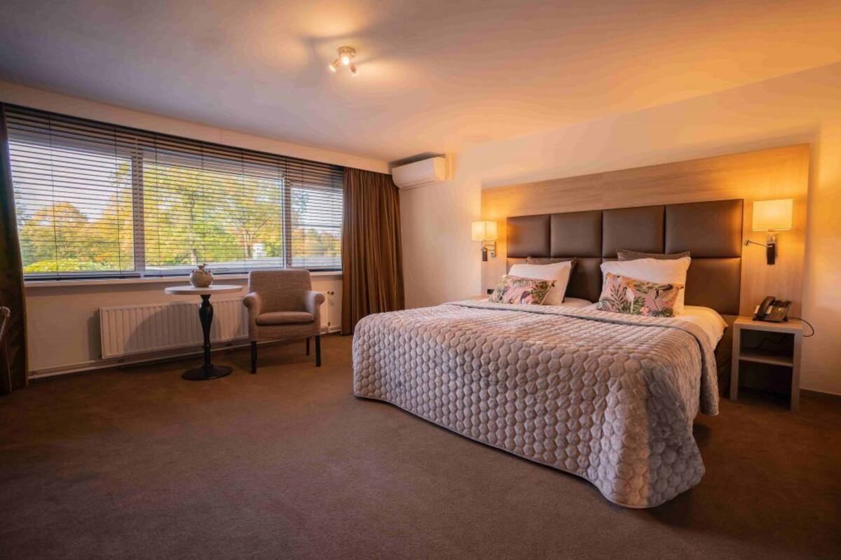 A hotel room with a large bed, a bedside table, a chair, and a window with blinds.