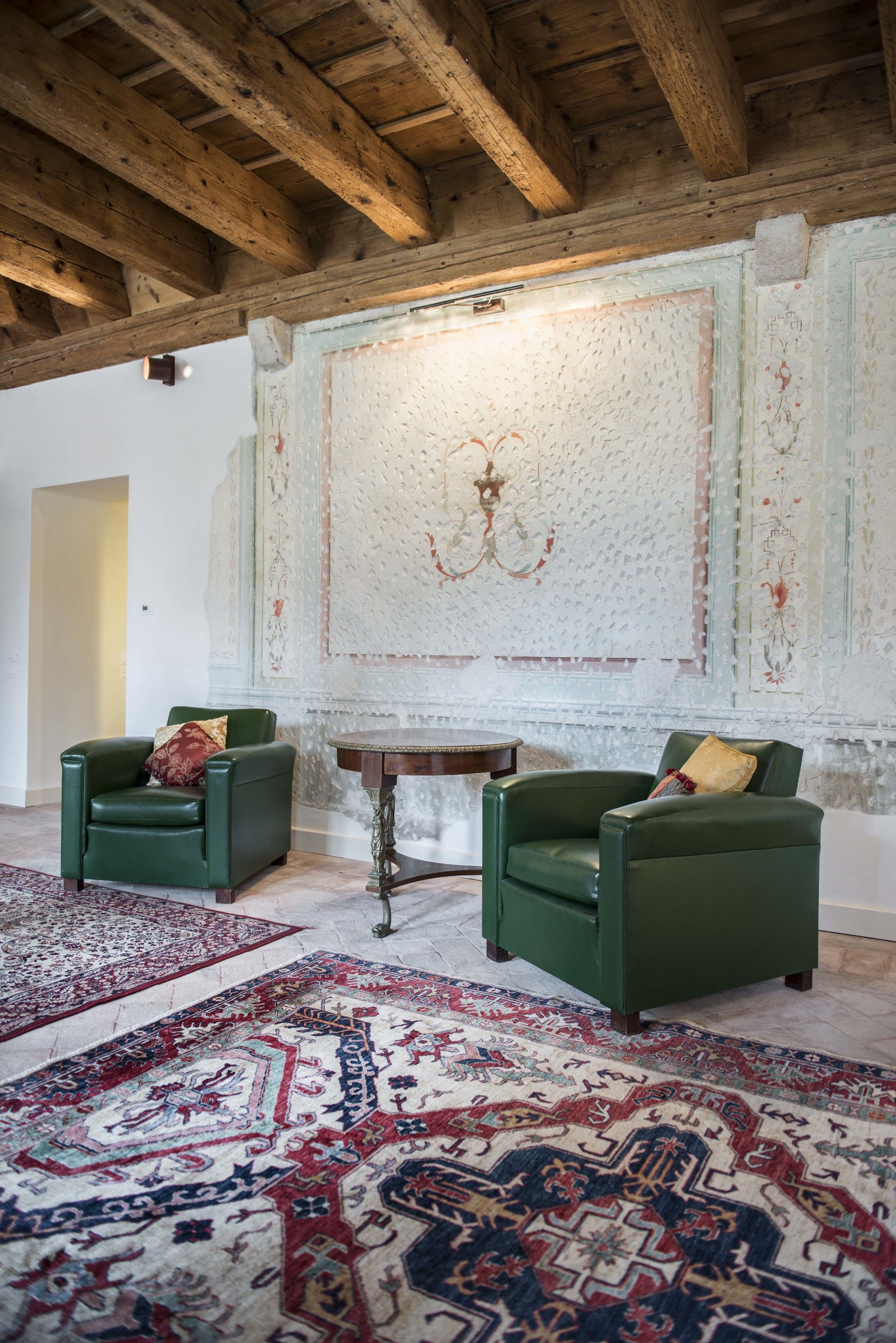 Een kamer met twee groene fauteuils, een ronde houten tafel en een groot, versierd wandtapijt.