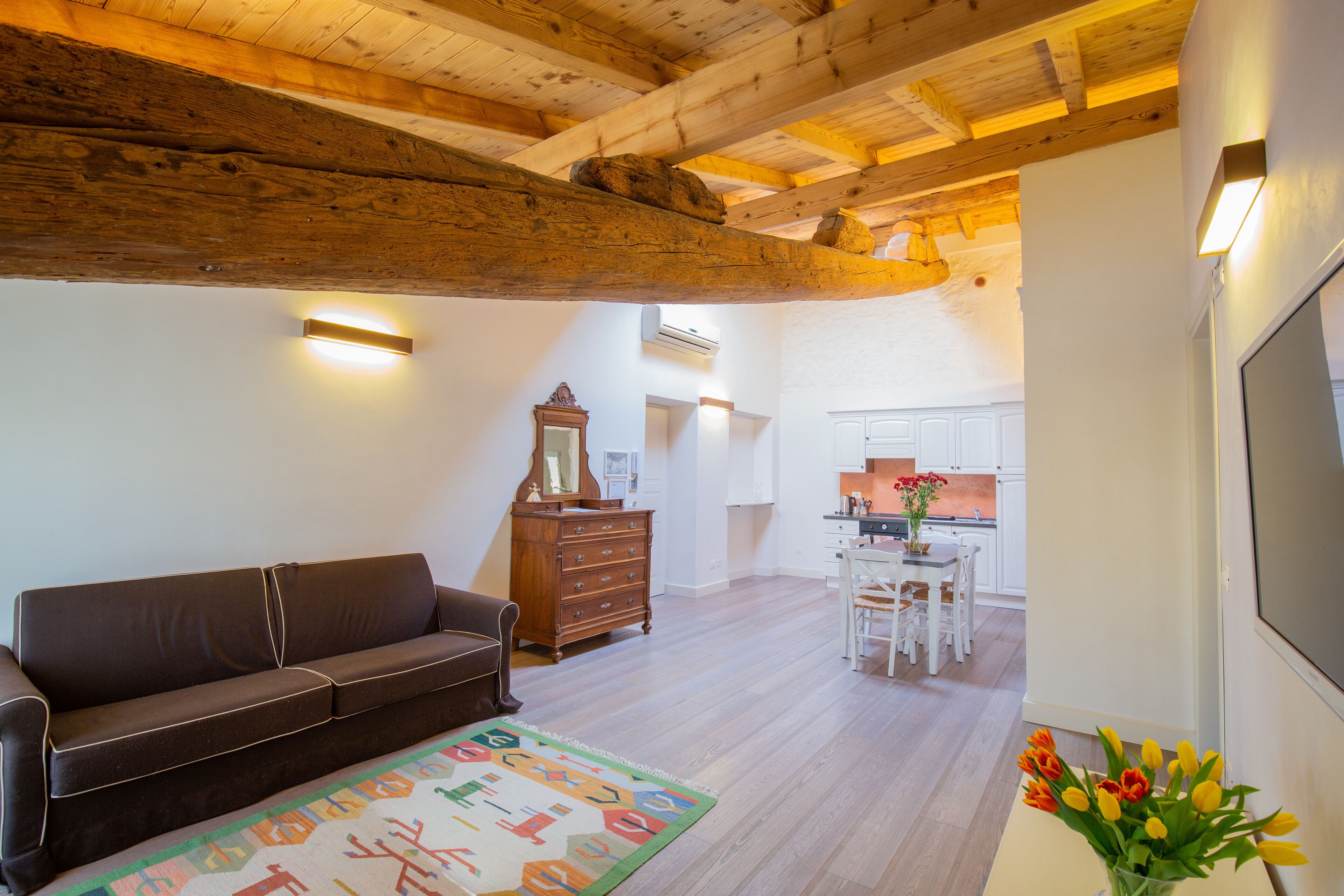 Un salon chaleureux avec un canapé en cuir marron, une commode en bois et un tapis coloré. On aperçoit à l’arrière-plan un coin repas avec des chaises blanches et une table.