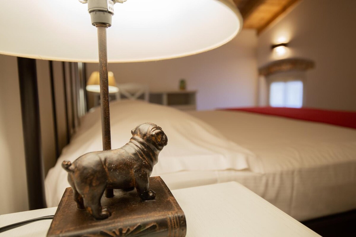 A bed with a white comforter and a red blanket, a lamp with a metal base, and a small metal sculpture of a dog on a wooden stand.