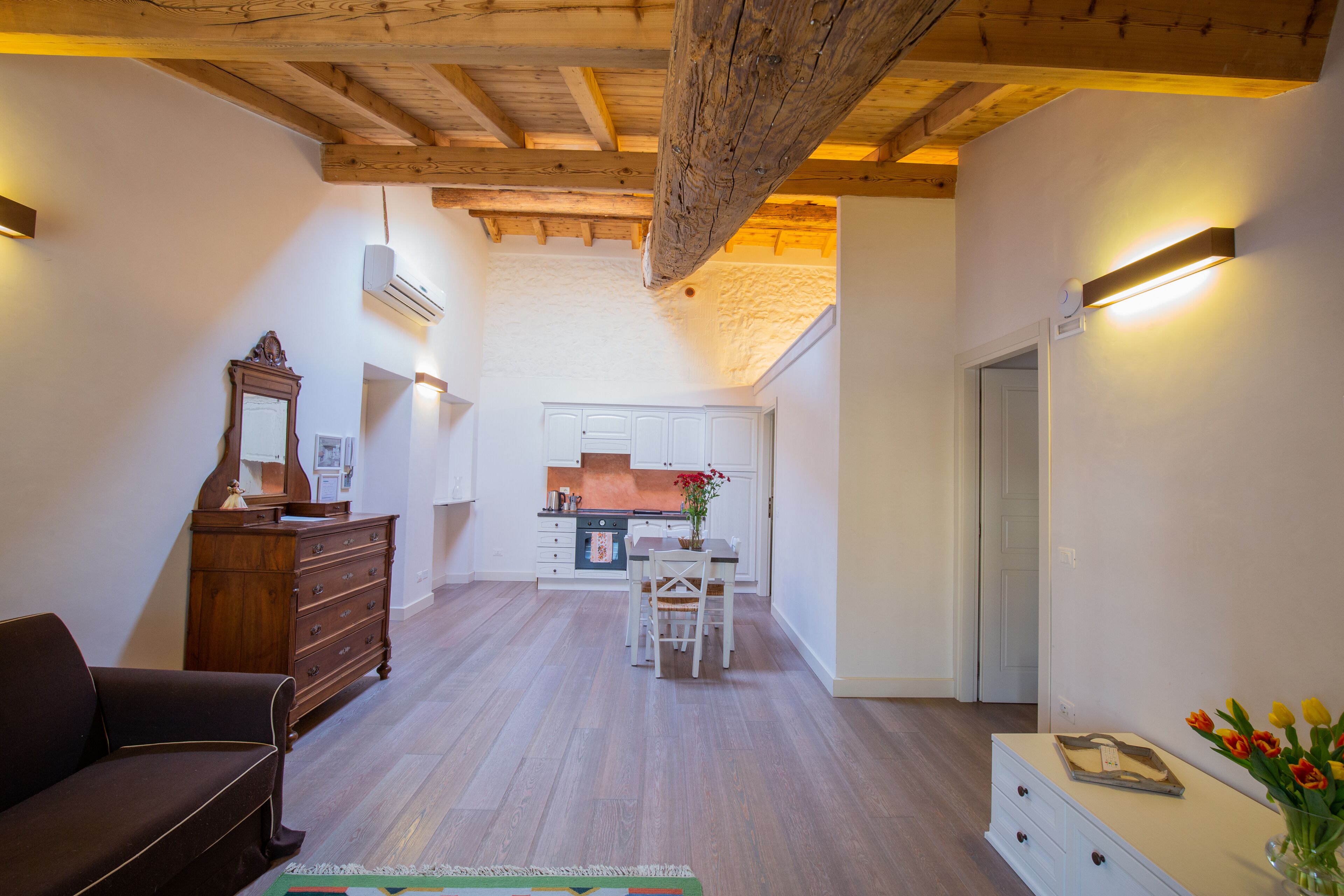 Uma sala de estar espaçosa com teto de madeira, uma mesa de jantar com cadeiras, um sofá, um aparador e um vaso com flores.