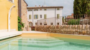 A swimming pool with a stone wall and a building with green shutters in the background. - Residence Corte San Carlo (Lazise)