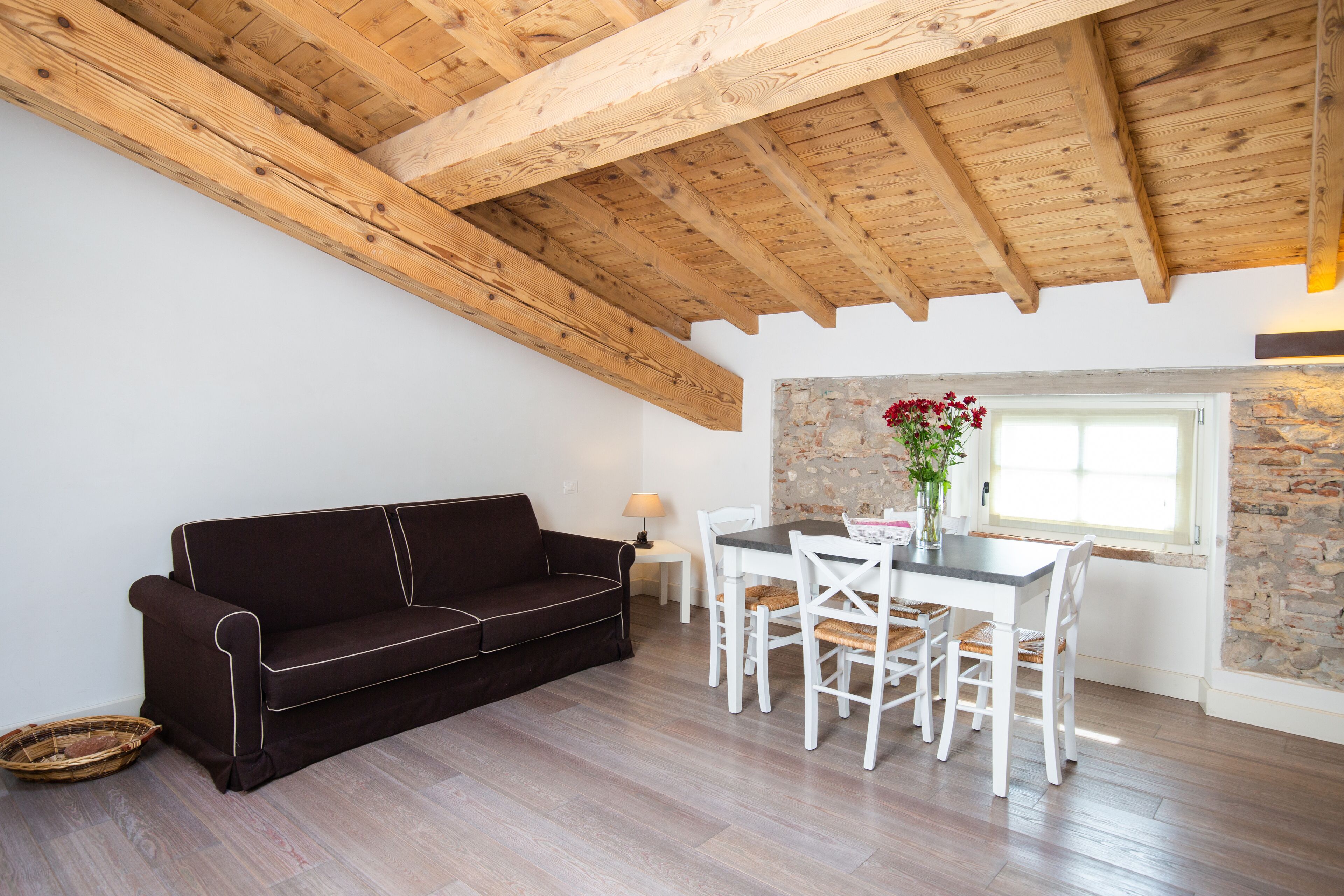Una stanza accogliente con soffitto in legno, un divano marrone scuro, un tavolo da pranzo bianco con sedie e un muro in pietra.